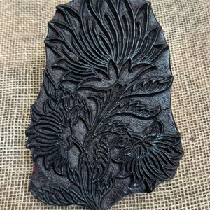 May include: A dark, rectangular wooden printing block featuring a detailed floral design. The block showcases a central flower with multiple petals, surrounded by leaves and smaller flowers. The intricate carving is visible against the dark background.