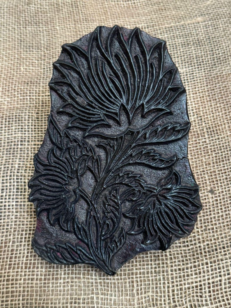 May include: A dark, rectangular wooden printing block featuring a detailed floral design. The block showcases a central flower with multiple petals, surrounded by leaves and smaller flowers. The intricate carving is visible against the dark background.