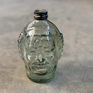 May include: A clear glass bottle shaped like a human face. The bottle has a black cap and a textured surface.