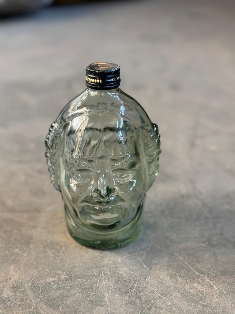 May include: A clear glass bottle shaped like a human face. The bottle has a black cap and a textured surface.