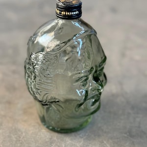 May include: A clear glass bottle shaped like a human head with a black cap. The bottle is green tinted and has a textured surface.