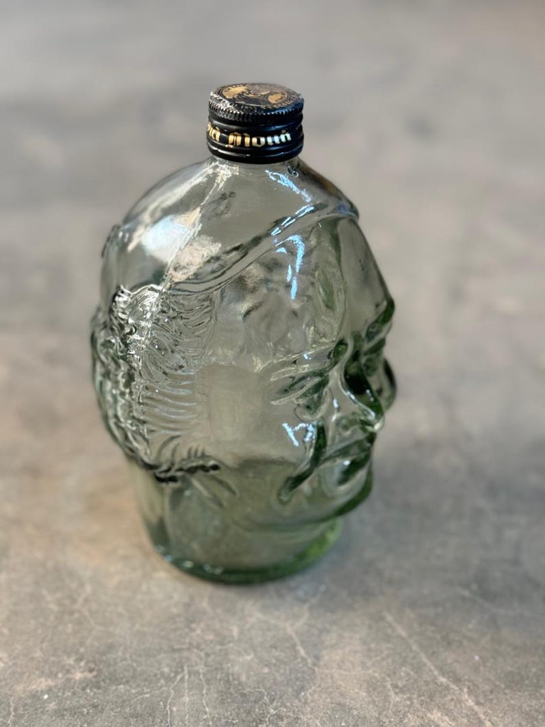 May include: A clear glass bottle shaped like a human head with a black cap. The bottle is green tinted and has a textured surface.