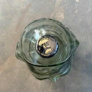 May include: A clear glass bottle shaped like a human skull with a black and gold sticker that reads "Mohan Meakin Limited, Est. 1855, For Sale Only".