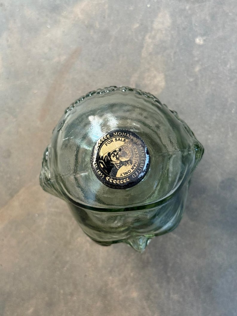 May include: A clear glass bottle shaped like a human skull with a black and gold sticker that reads "Mohan Meakin Limited, Est. 1855, For Sale Only".