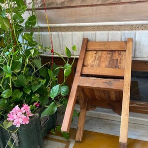 Puede incluir: Una silla de madera con un acabado marr&oacute;n est&aacute; tumbada de lado en un suelo de baldosas. La silla tiene un respaldo y un asiento de listones. Una planta en maceta con flores rosas est&aacute; en el fondo.