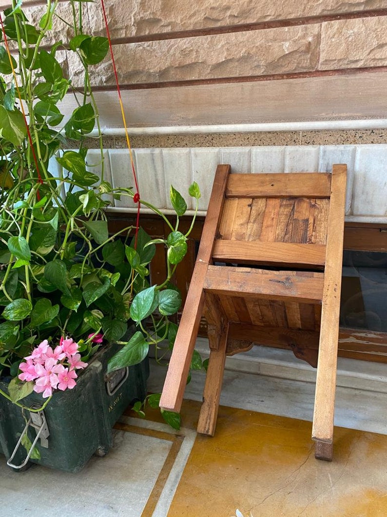 Puede incluir: Una silla de madera con un acabado marr&oacute;n est&aacute; tumbada de lado en un suelo de baldosas. La silla tiene un respaldo y un asiento de listones. Una planta en maceta con flores rosas est&aacute; en el fondo.