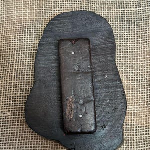 May include: A dark brown, irregularly shaped wooden stamp with a rectangular handle attached. The stamp has a rough, textured surface and appears to be handmade. Small, shiny embellishments are visible on the handle.