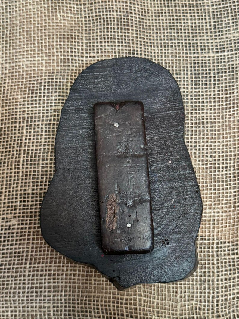 May include: A dark brown, irregularly shaped wooden stamp with a rectangular handle attached. The stamp has a rough, textured surface and appears to be handmade. Small, shiny embellishments are visible on the handle.