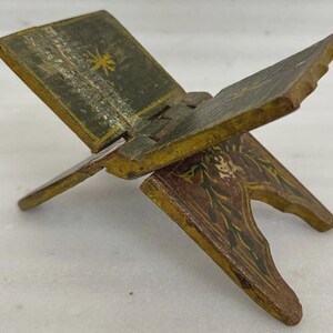 Puede incluir: Un peque&ntilde;o atril de madera antiguo con dise&ntilde;o de bisagra. Pintado en tonos verdes, amarillos y marrones, con motivos florales y de estrella. Dise&ntilde;ado para sostener un libro abierto, mide aproximadamente 15 cm de largo.