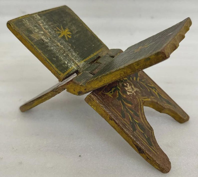 Puede incluir: Un peque&ntilde;o atril de madera antiguo con dise&ntilde;o de bisagra. Pintado en tonos verdes, amarillos y marrones, con motivos florales y de estrella. Dise&ntilde;ado para sostener un libro abierto, mide aproximadamente 15 cm de largo.