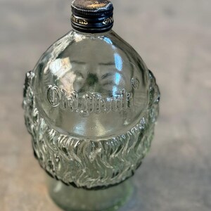 May include: A clear glass bottle with a textured surface and a black cap. The bottle is shaped like a rounded rectangle and has the words "Old Grand-Dad" embossed on the front.