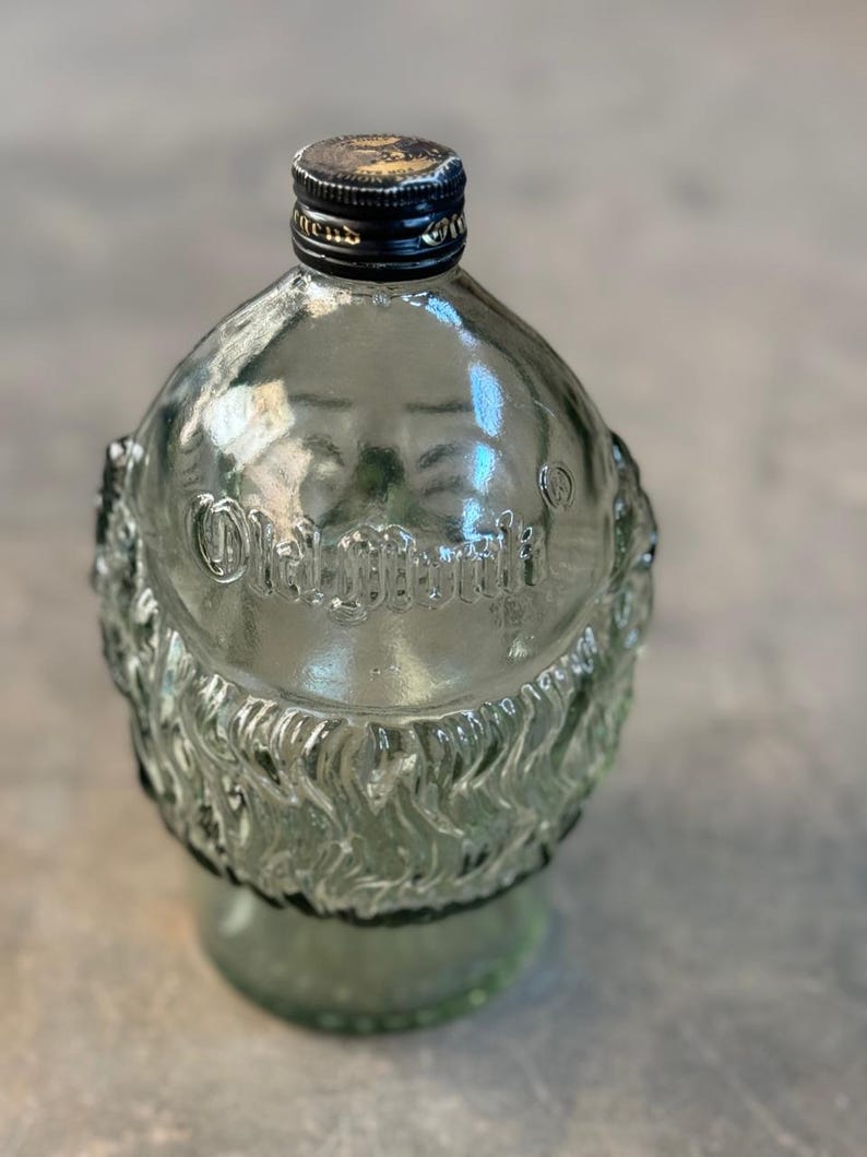 May include: A clear glass bottle with a textured surface and a black cap. The bottle is shaped like a rounded rectangle and has the words "Old Grand-Dad" embossed on the front.