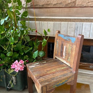 Puede incluir: Una silla de madera r&uacute;stica con un acabado desgastado multicolor. La silla tiene un dise&ntilde;o sencillo con un asiento rectangular y un respaldo hecho de tablones de madera recuperados. La silla est&aacute; sentada en un suelo de baldosas con una planta en maceta y flores blancas en primer plano.