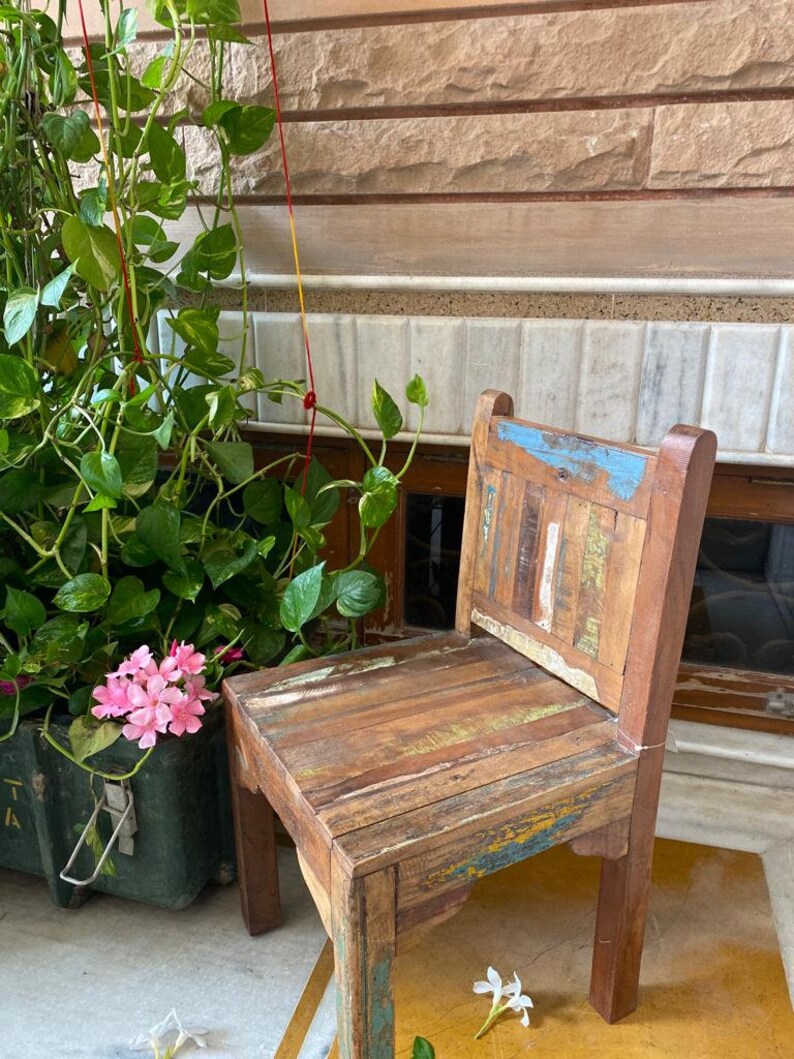 Puede incluir: Una silla de madera r&uacute;stica con un acabado desgastado multicolor. La silla tiene un dise&ntilde;o sencillo con un asiento rectangular y un respaldo hecho de tablones de madera recuperados. La silla est&aacute; sentada en un suelo de baldosas con una planta en maceta y flores blancas en primer plano.