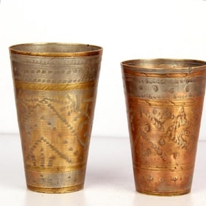 May include: Two tapered metal tumblers featuring a mix of brass and copper tones. The surfaces are decorated with horizontal bands of etched geometric patterns, including small dots, diagonal lines, and triangular shapes around the exterior.