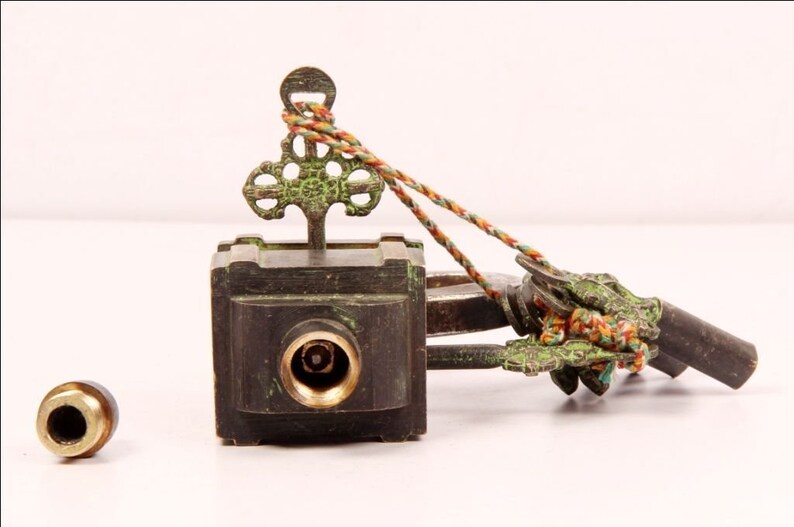 May include: A brass key with an ornate design and a green patina. The key is attached to a small, square box with a keyhole. The box is also made of brass and has a green patina. The key and box are connected by a string.