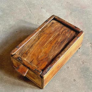 May include: A rectangular wooden box with a natural brown finish and visible wood grain. The box features a sliding lid mechanism with a small protruding tab on one end and reinforced edges secured with small metal nails along the frame.