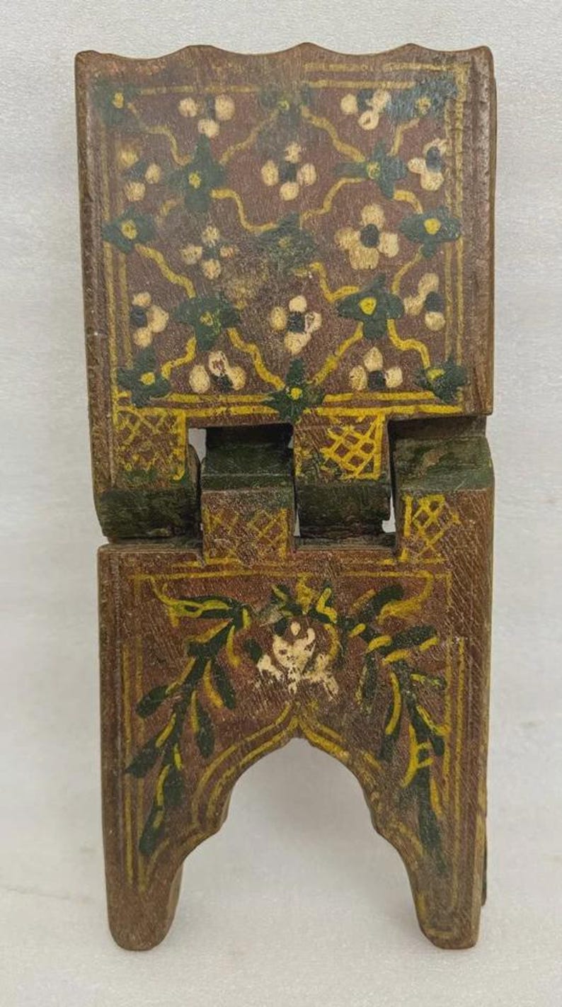 Puede incluir: Un peque&ntilde;o soporte de madera plegable con base marr&oacute;n y dise&ntilde;os florales intrincados en blanco, verde y amarillo. El soporte est&aacute; pintado con un patr&oacute;n detallado y tiene un arco decorativo en la base.