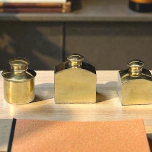 May include: Three brass inkwells of different shapes are arranged on a wooden desk, with a pen and a leather notepad. The inkwells have a vintage appearance, with a cylindrical, a rectangular, and a square design. A potted plant is visible in the background.