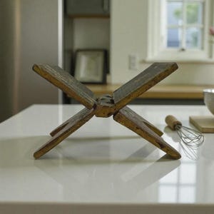 Puede incluir: Un soporte de libro de cocina r&uacute;stico de madera en forma de X, con un aspecto envejecido y desgastado. El soporte est&aacute; hecho de varias piezas de madera unidas. Un batidor y un cuenco est&aacute;n en el fondo.