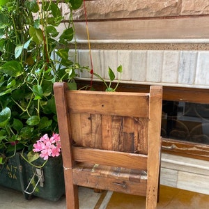 Puede incluir: Una silla de madera con respaldo y asiento de listones. La silla es marr&oacute;n y tiene un aspecto r&uacute;stico. Est&aacute; situada delante de una pared de ladrillo con una planta verde en una maceta.