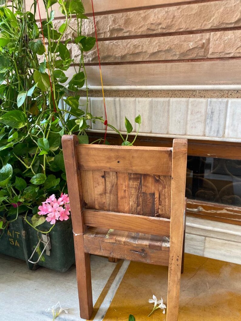 Puede incluir: Una silla de madera con respaldo y asiento de listones. La silla es marr&oacute;n y tiene un aspecto r&uacute;stico. Est&aacute; situada delante de una pared de ladrillo con una planta verde en una maceta.