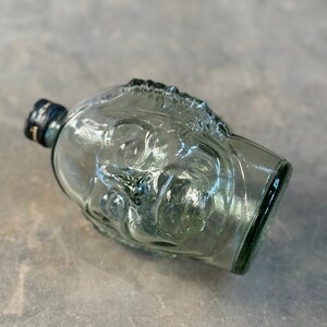 May include: A clear glass bottle shaped like a human head. The bottle has a textured surface and a dark green cap.