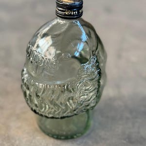 May include: A clear glass bottle shaped like a Santa Claus head with a black screw-on lid. The bottle is textured and has a white label with the word "Old" on it.
