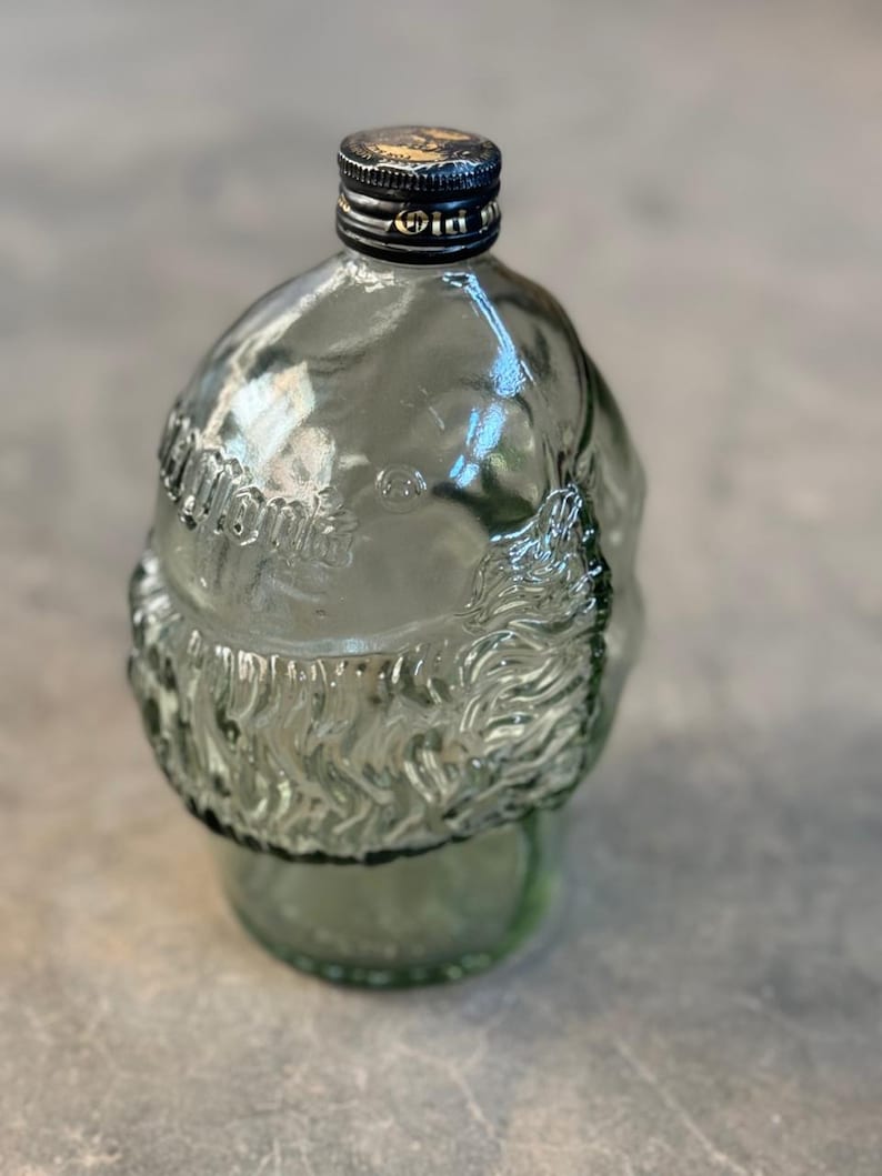May include: A clear glass bottle shaped like a Santa Claus head with a black screw-on lid. The bottle is textured and has a white label with the word "Old" on it.