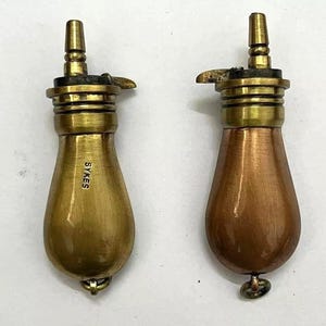 以下が含まれることがあります： 真鍮製のキャップとノズルが付いた、洋梨型の火薬入れ2点。左側の火薬入れは真鍮製で、本体に「SYKES」の刻印があります。右側の火薬入れは銅製の本体に真鍮製のキャップが付いています。どちらも底面に小さな金属製のリングが付いています。