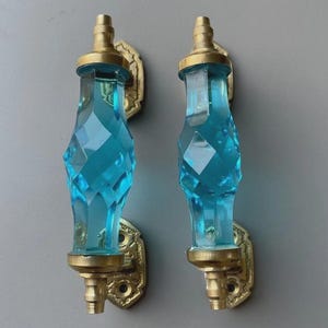 Puede incluir: Dos manijas de puerta verticales con centros de vidrio azul turquesa translúcido facetado con un patrón de corte de diamante. Cada manija está asegurada por placas de montaje de latón cepillado ornamentadas, terminadas con pequeños remates cilíndricos.