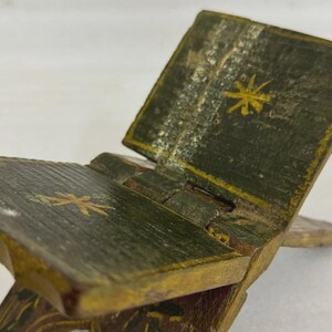Puede incluir: Un peque&ntilde;o atril plegable de madera pintado en tonos verdes y dorados. El atril presenta un dise&ntilde;o con bisagras y est&aacute; decorado con motivos dorados de estrellas. La apariencia envejecida sugiere un art&iacute;culo vintage o antiguo.