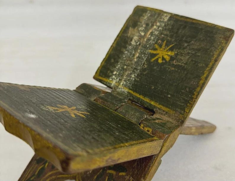 Puede incluir: Un peque&ntilde;o atril plegable de madera pintado en tonos verdes y dorados. El atril presenta un dise&ntilde;o con bisagras y est&aacute; decorado con motivos dorados de estrellas. La apariencia envejecida sugiere un art&iacute;culo vintage o antiguo.