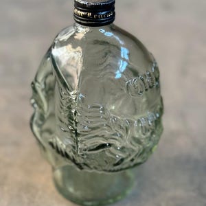 May include: A clear glass bottle with a textured surface and a black cap. The bottle is shaped like a head and has a design of a face on the front.