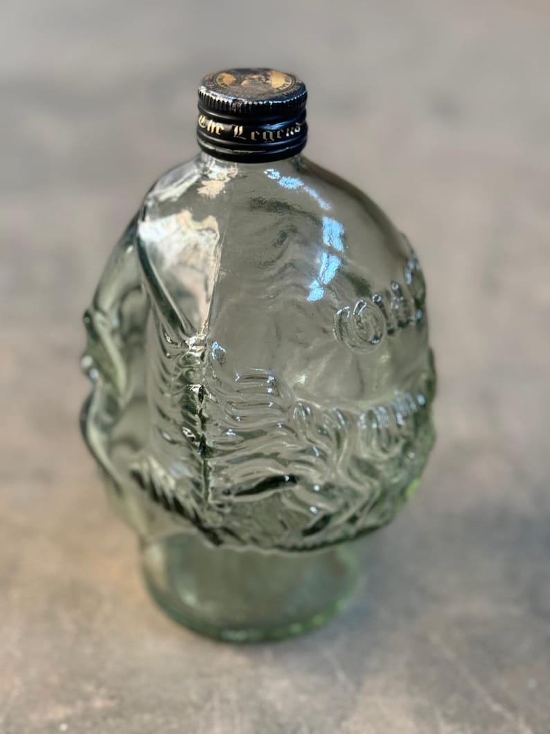 May include: A clear glass bottle with a textured surface and a black cap. The bottle is shaped like a head and has a design of a face on the front.
