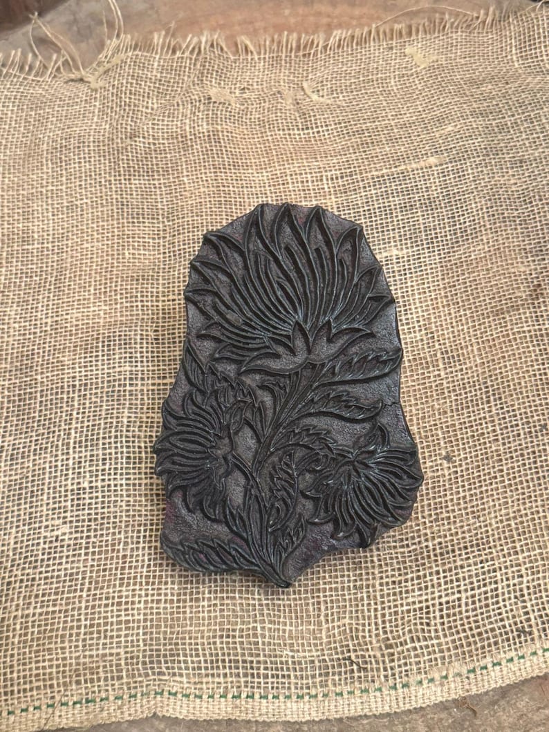 May include: A dark brown, rectangular wooden block with a floral design. The carved pattern features a large central flower with detailed petals and leaves, along with smaller flowers. The block is set on a rough, tan burlap surface.