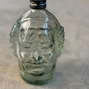 May include: A clear glass bottle shaped like a human face. The bottle has a black lid with a gold band.