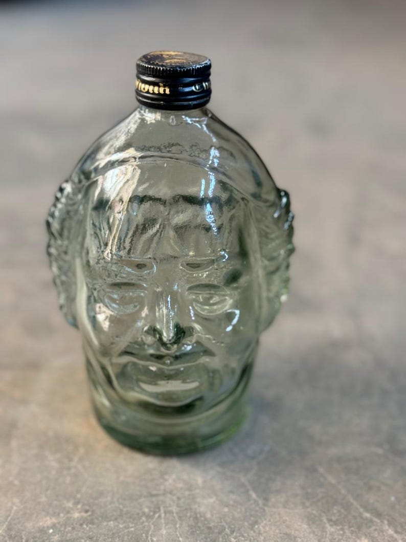 May include: A clear glass bottle shaped like a human face. The bottle has a black lid with a gold band.