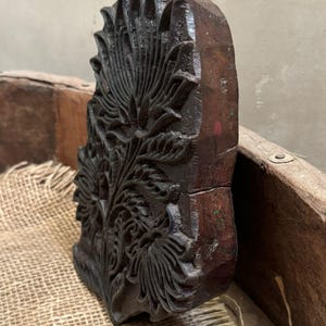May include: A dark brown, intricately carved wooden block featuring a floral design. The block has a rectangular shape with a detailed pattern of leaves and petals. The wood has a rich, aged appearance, suggesting it is vintage or antique.