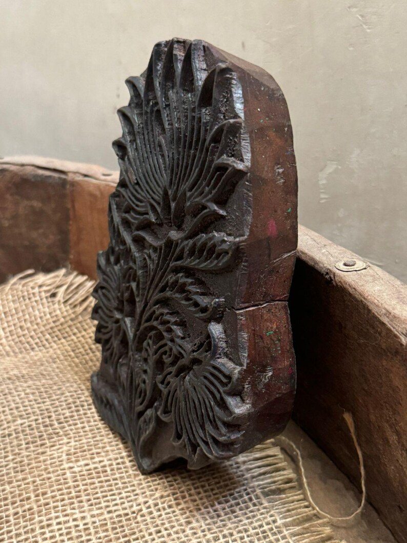 May include: A dark brown, intricately carved wooden block featuring a floral design. The block has a rectangular shape with a detailed pattern of leaves and petals. The wood has a rich, aged appearance, suggesting it is vintage or antique.