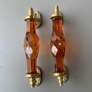 May include: Two matching door handles featuring faceted, translucent amber-colored glass centers with a diamond-cut texture. Each handle is secured by polished brass hardware at the top and bottom, including decorative mounting plates with screw holes.