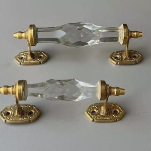 May include: A pair of clear crystal door handles with faceted designs. They are mounted on ornate, gold-coloured metal bases. The hardware has a vintage aesthetic, suitable for interior design projects.