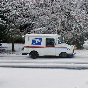 May include: A white Royal Mail delivery van driving on a snow-covered road. The van has the Royal Mail logo on the side and is surrounded by snow-covered trees. The road is partially cleared.