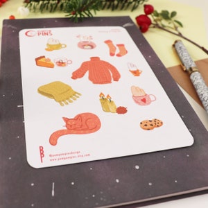 Op de afbeelding: Een vel stickers met illustraties van knusse winterartikelen, waaronder een kat, een mok, een trui, kaarsen en koekjes. De stickers zijn op een witte achtergrond en hebben een handschrift.