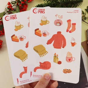 Op de afbeelding: Twee vellen stickers met een knus winterthema. De stickers bevatten illustraties van een kat, een mok warme chocolademelk, een gebreide trui, een stuk taart, een kaars, een sjaal en meer. De stickers zijn gelabeld met "Pompom Pins" en "Lees- en knusse set".