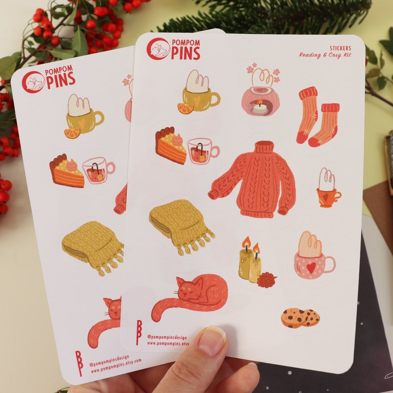 Op de afbeelding: Twee vellen stickers met een knus winterthema. De stickers bevatten illustraties van een kat, een mok warme chocolademelk, een gebreide trui, een stuk taart, een kaars, een sjaal en meer. De stickers zijn gelabeld met "Pompom Pins" en "Lees- en knusse set".