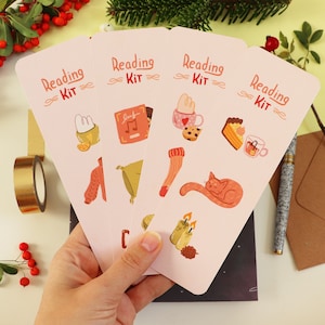 Peut inclure: Quatre marque-pages roses avec le texte "Reading Kit" et diverses illustrations, dont un chat, des bougies, un livre et une tasse de café.