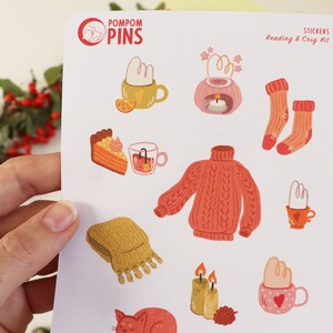 Op de afbeelding: Een vel stickers met een knus winterthema. De stickers bevatten illustraties van een warme trui, een mok met warme chocolademelk, een stuk taart, een kaars, een kat en een sjaal. De stickers zijn perfect om een ​​beetje warmte en comfort toe te voegen aan je planner, dagboek of laptop.