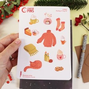 Op de afbeelding: Een vel stickers met illustraties van knusse winterartikelen, waaronder een trui, een mok warme chocolademelk, een kat en kaarsen. De stickers maken deel uit van een "Lees & Gezelligheidsset" en zijn van het merk "Pompom Pins".