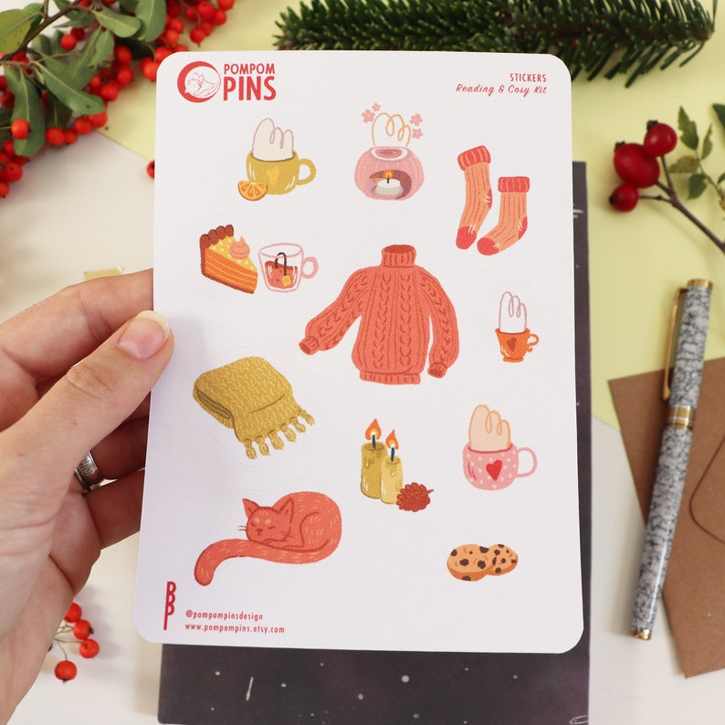 Op de afbeelding: Een vel stickers met illustraties van knusse winterartikelen, waaronder een trui, een mok warme chocolademelk, een kat en kaarsen. De stickers maken deel uit van een "Lees & Gezelligheidsset" en zijn van het merk "Pompom Pins".